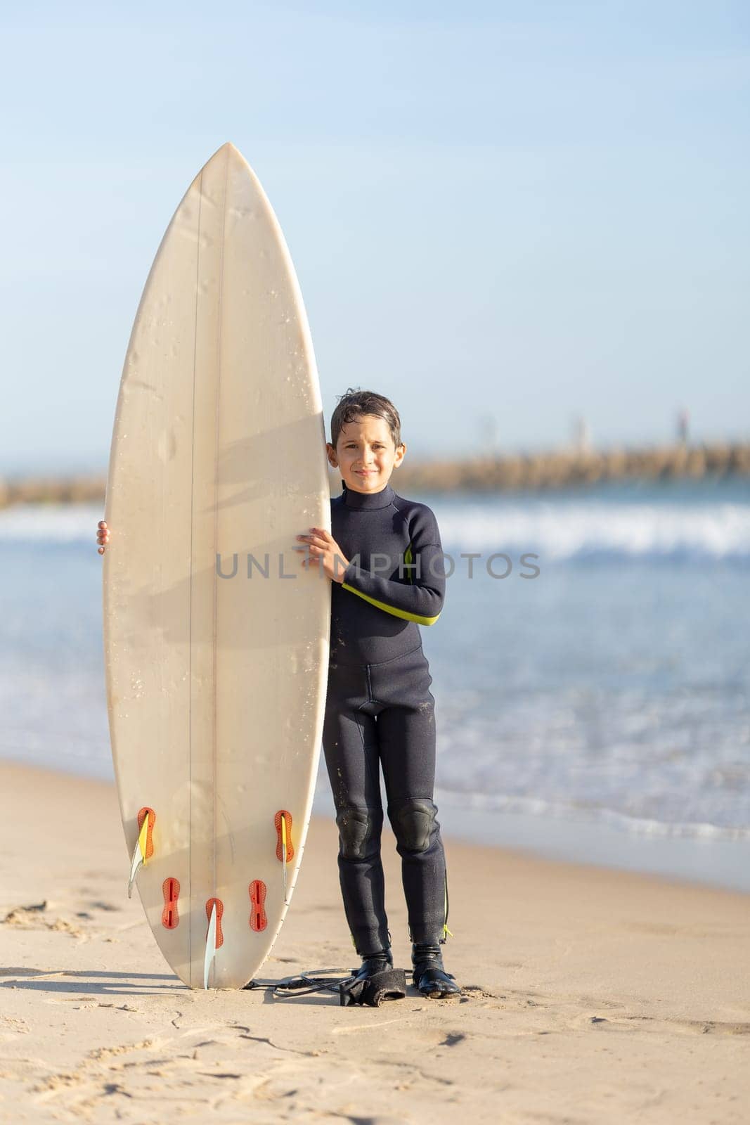 A little smiling boy standing on the seashore with a big surfing board. Vertical shot