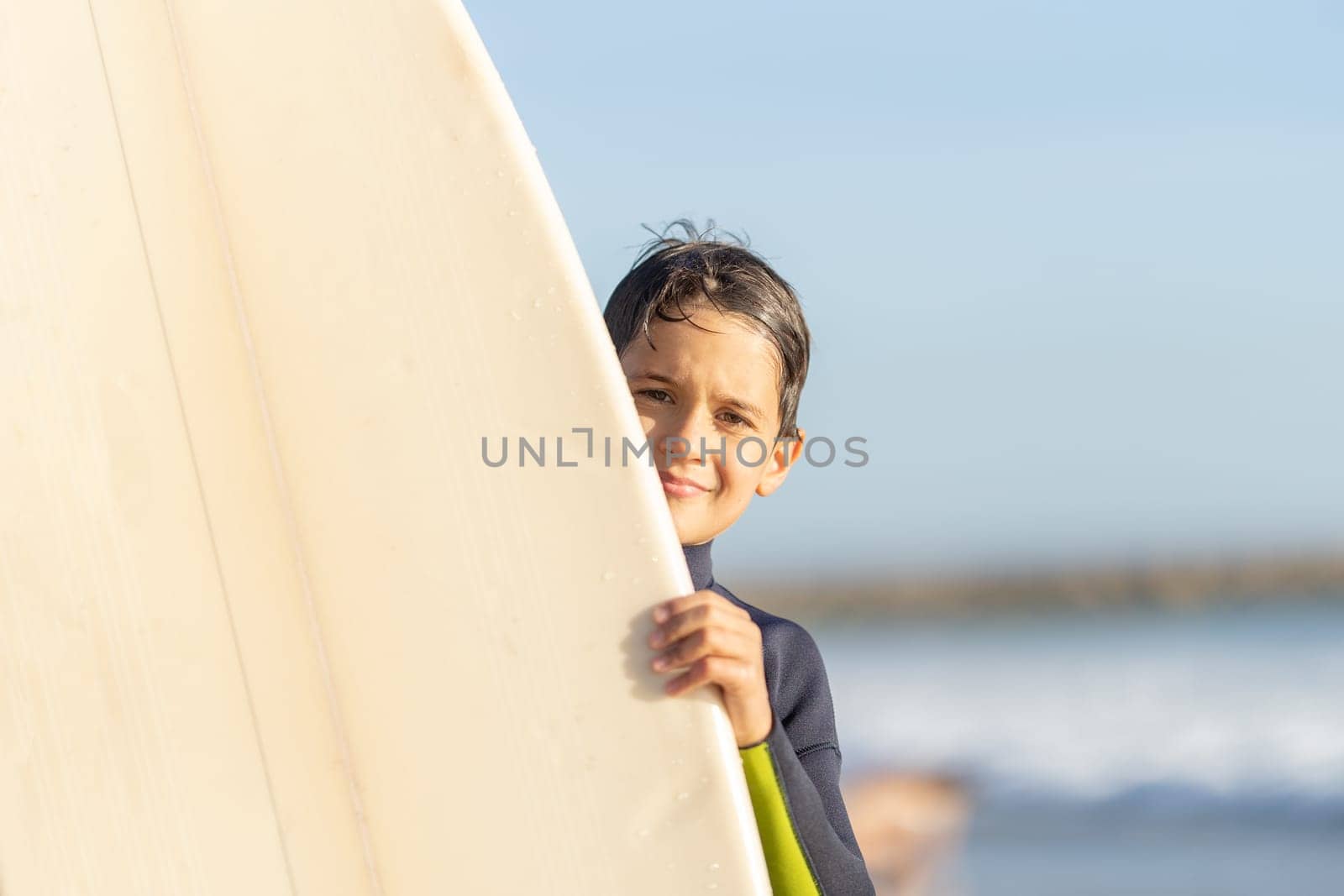 Little boy peeking out from behind a surfboard. Mid shot