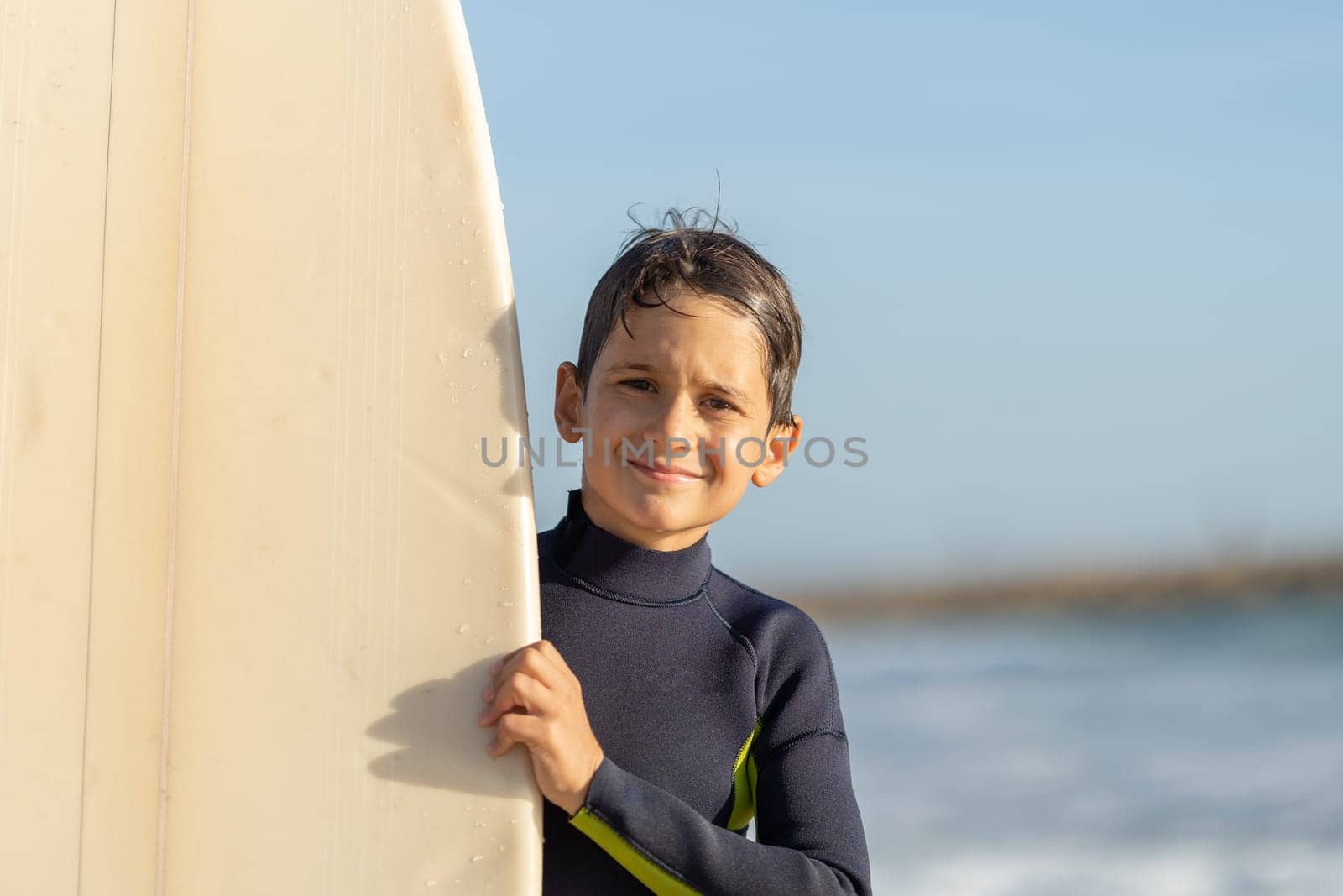 Smiling little boy peeking out from behind a surfboard. Mid shot