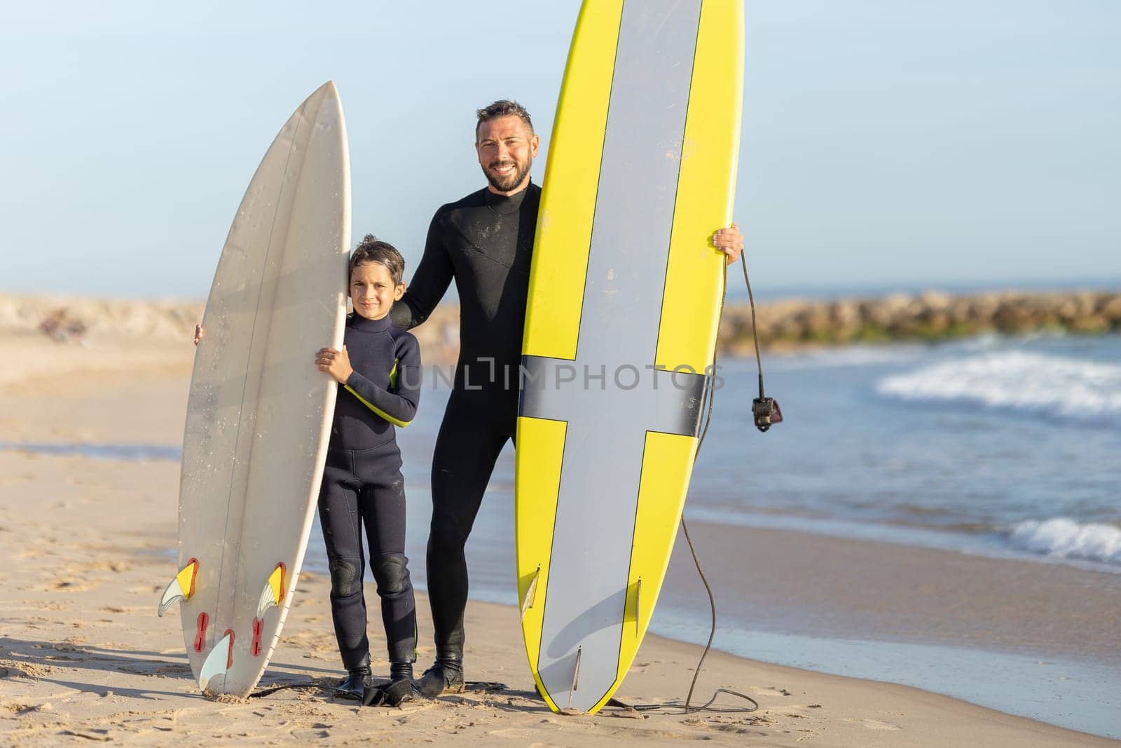 A family of surfers - smiling father and son standing on the seashore with their surfing boards. Mid shot