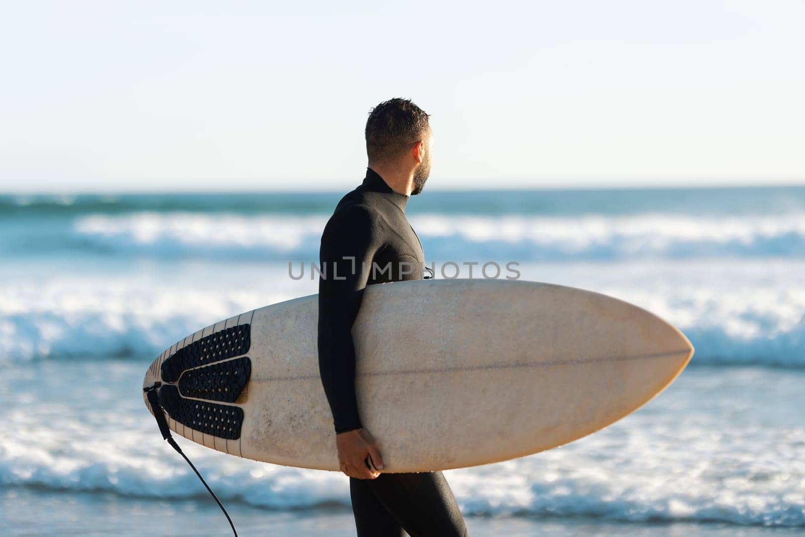 A man surfer in a wetsuit walking on the shore holding a surfboard and looking at the sea. Mid shot