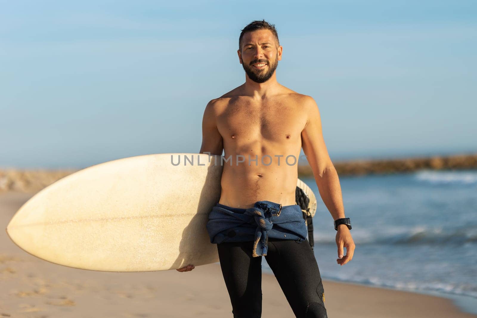 A man surfer with naked torso standing on the shore holding a surfing board. Mid shot