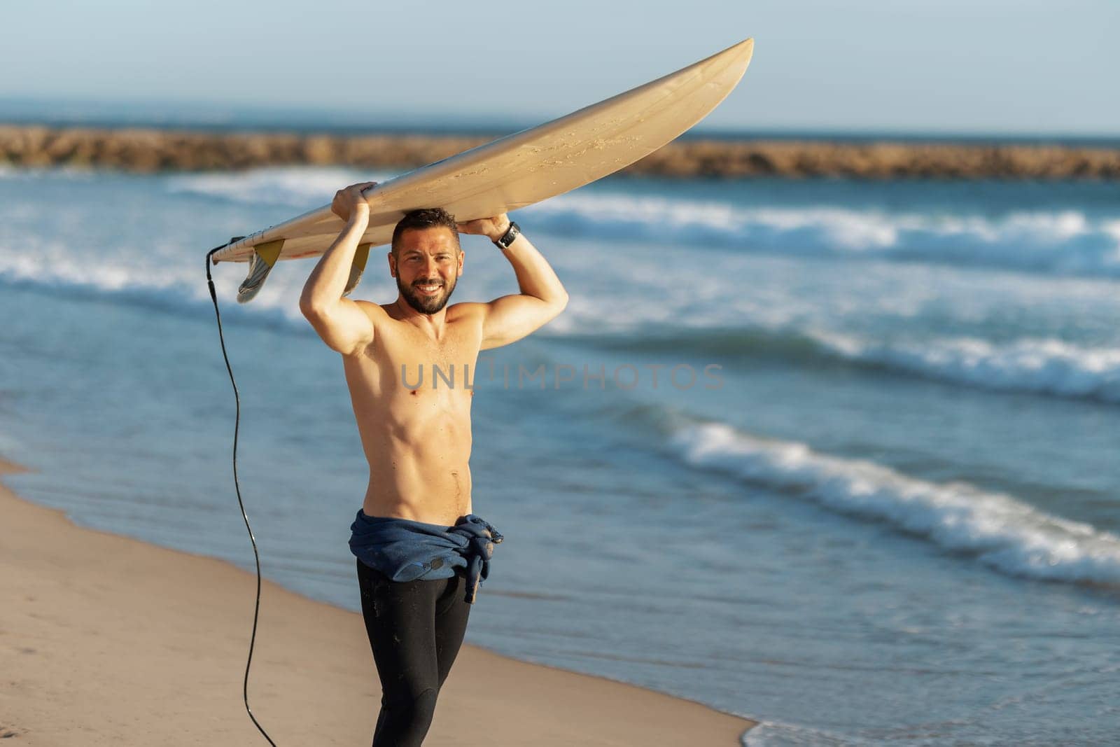 A smiling man surfer with naked torso holding a surfing board over his head. Mid shot