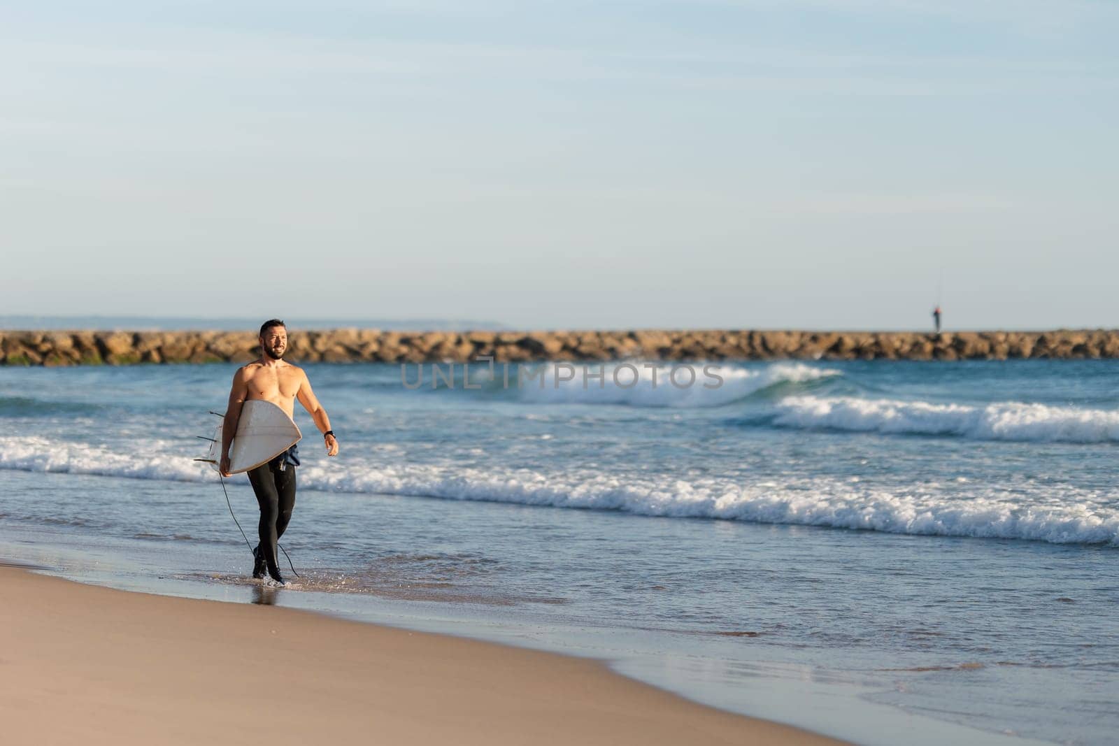 A man surfer with naked torso walking on the seashore. Mid shot