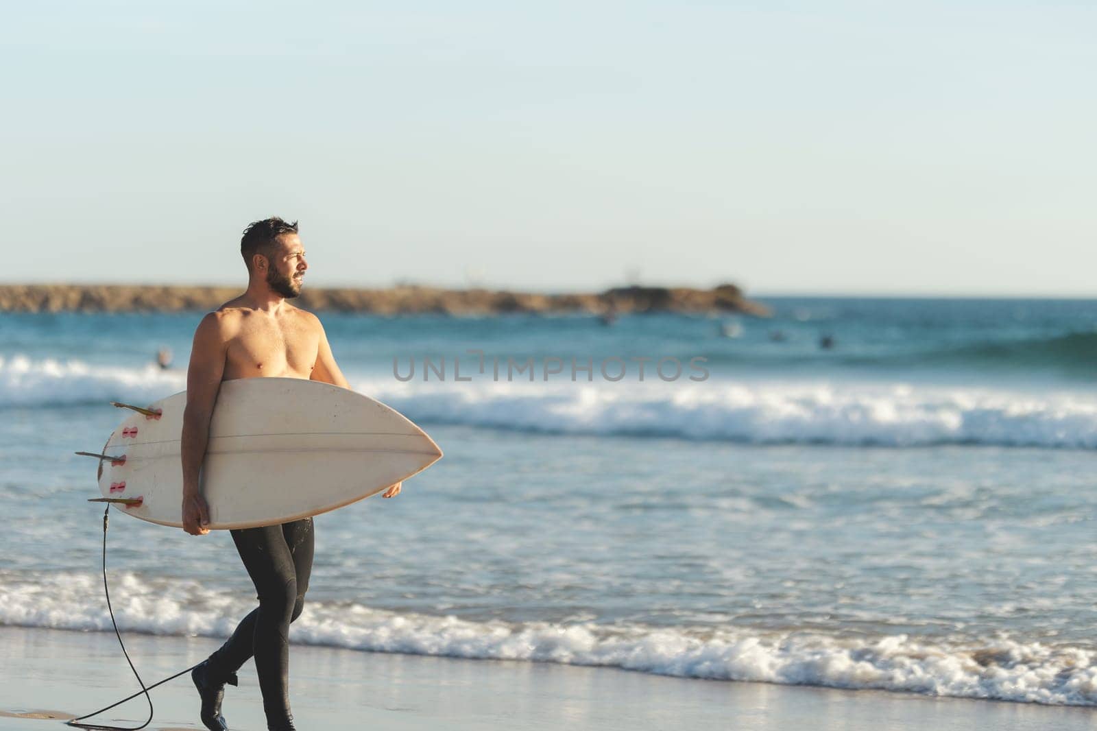 A smiling man surfer with naked torso walking on the seashore. Mid shot
