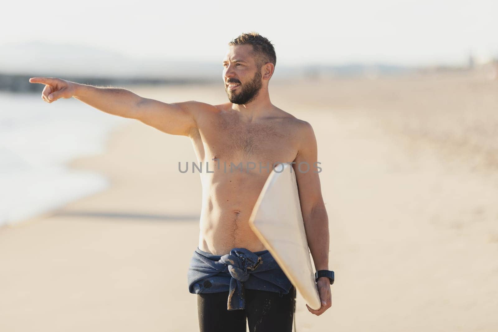 A man surfer with naked torso pointing at the sea. Mid shot