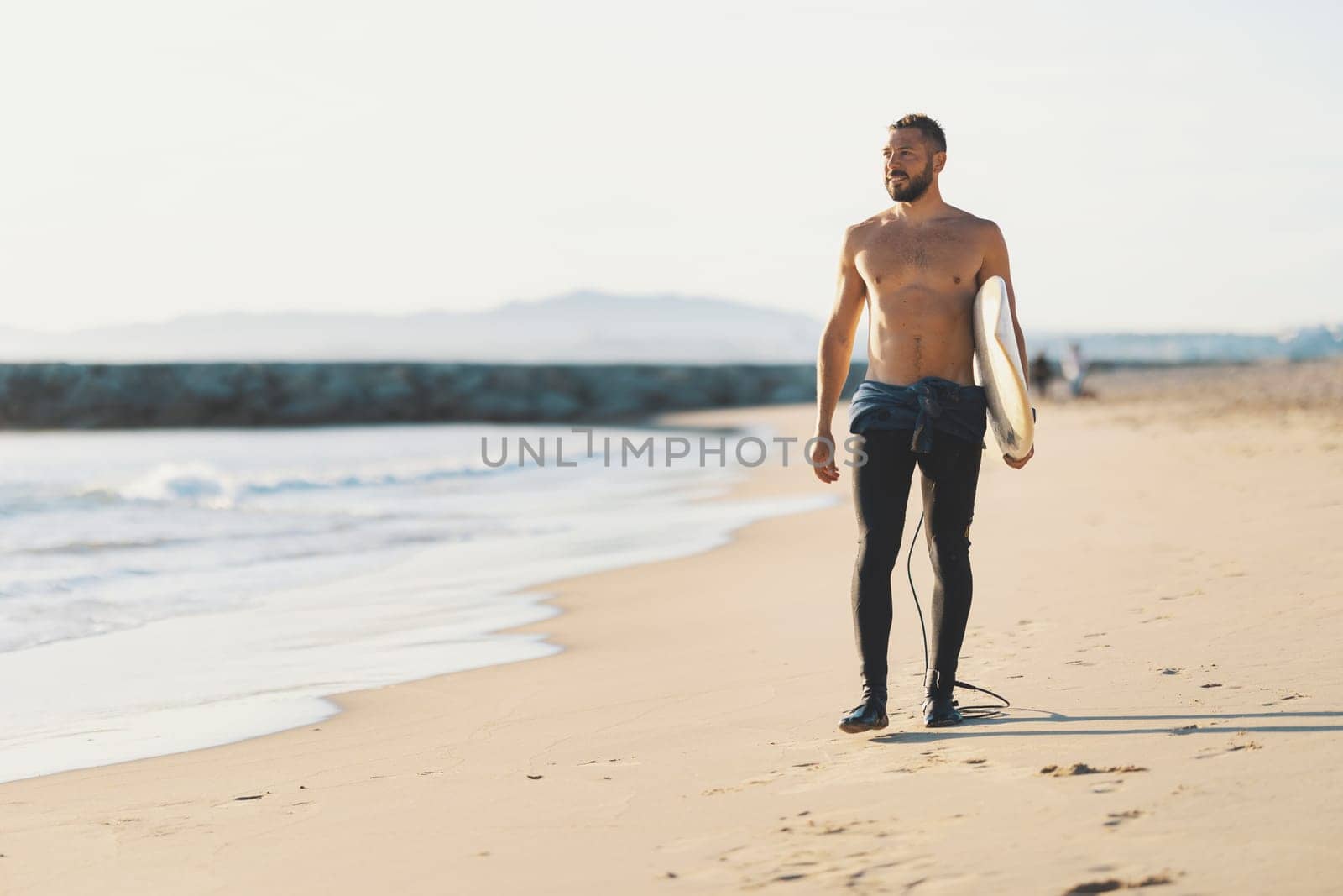 An athletic man surfer with naked torso standing on the seashore. Mid shot