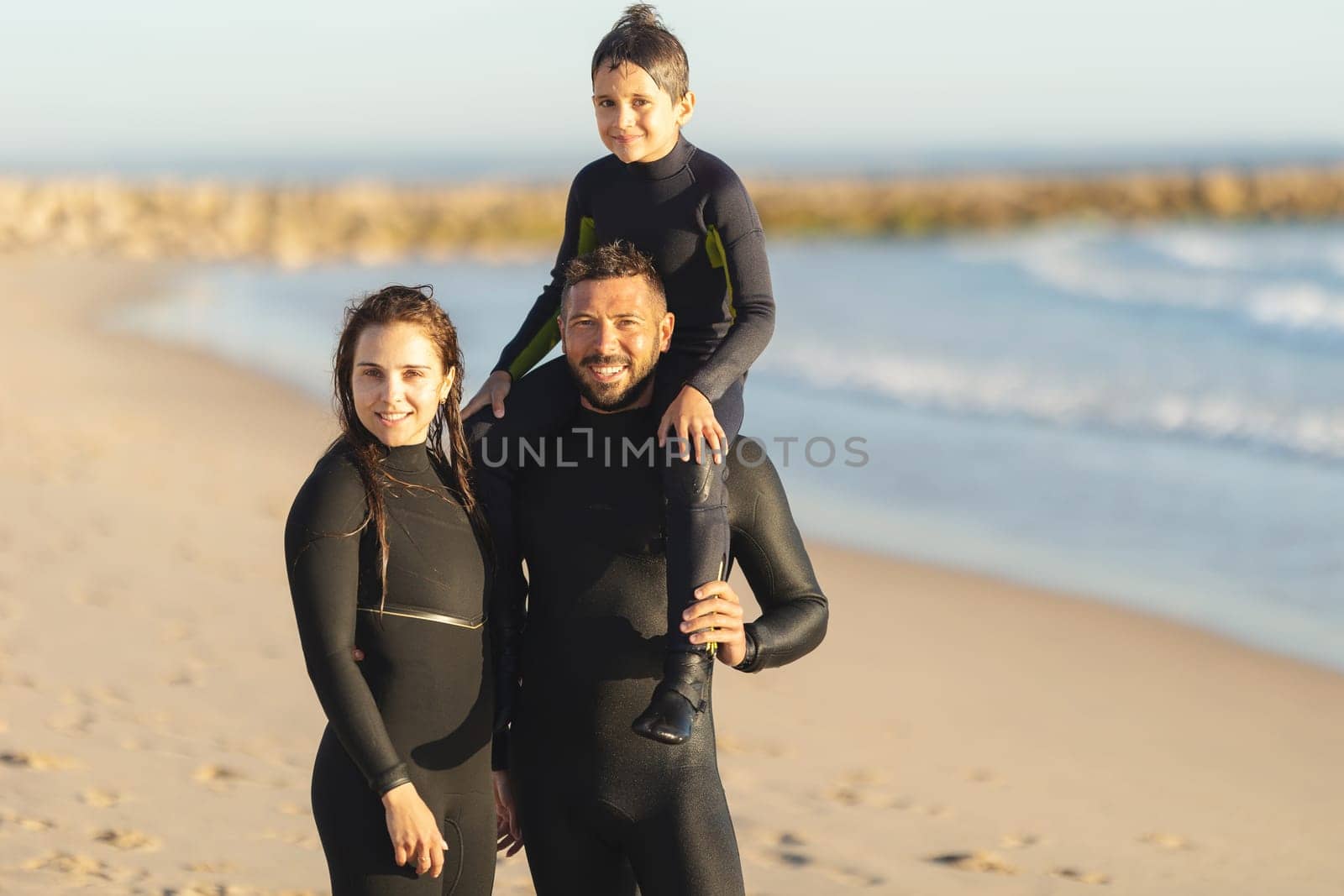 A family of surfers stands on the seashore in wetsuits by Studia72