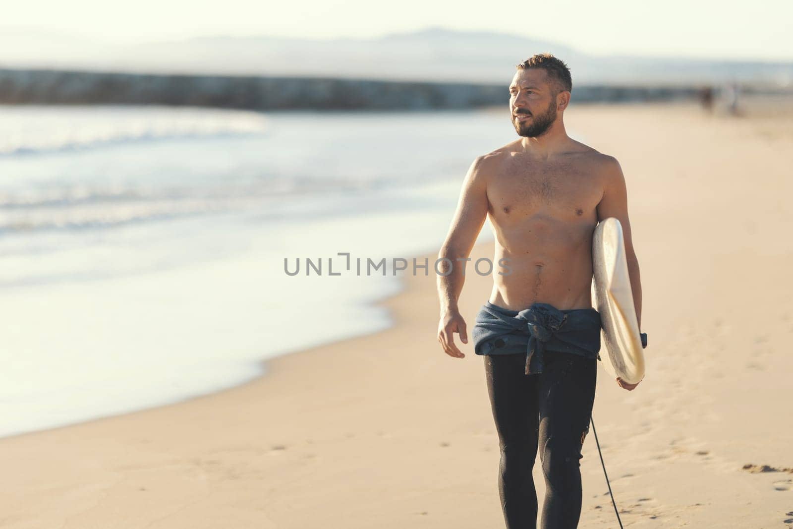 An athletic man surfer with naked torso walking on the seashore. Mid shot