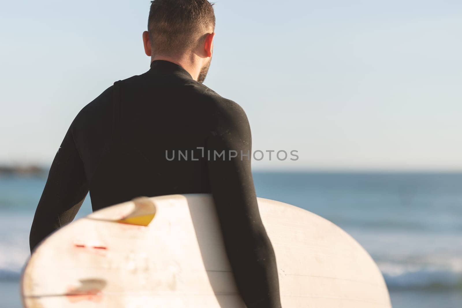 An athletic man surfer in a wetsuit standing by the ocean. Mid shot