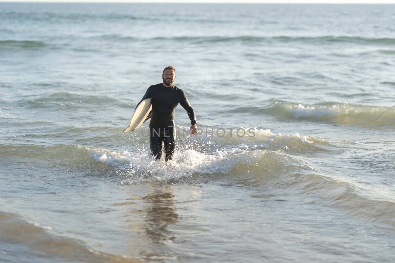 Smiling man surfer coming out of the sea holding a surfboard. Mid shot