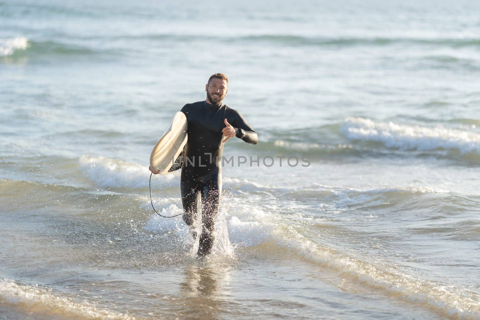 Smiling man surfer running out of the sea holding a surfboard and showing shaka. Mid shot