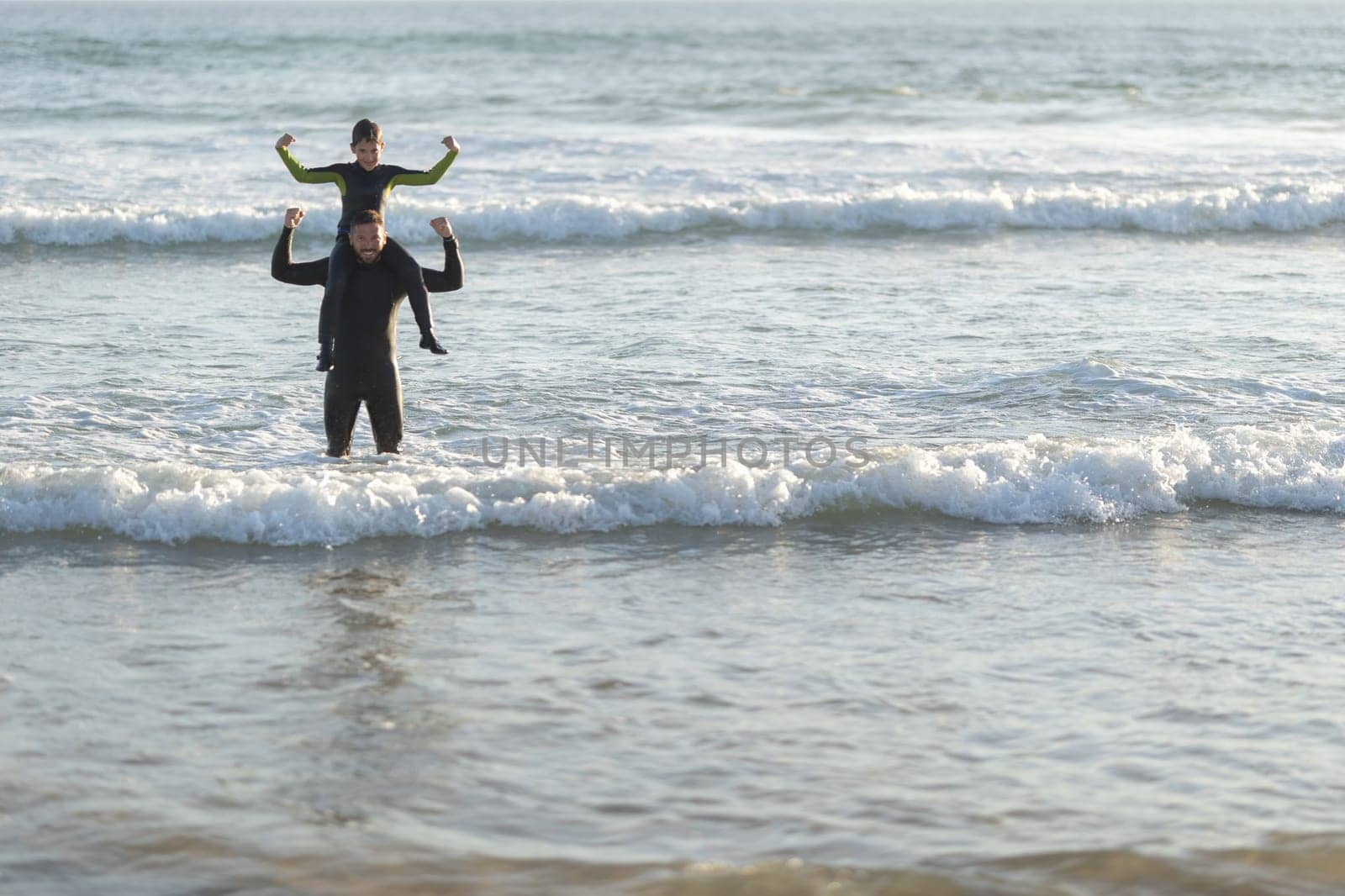 Little son on the shoulders of his father in the sea showing muscles. Mid shot