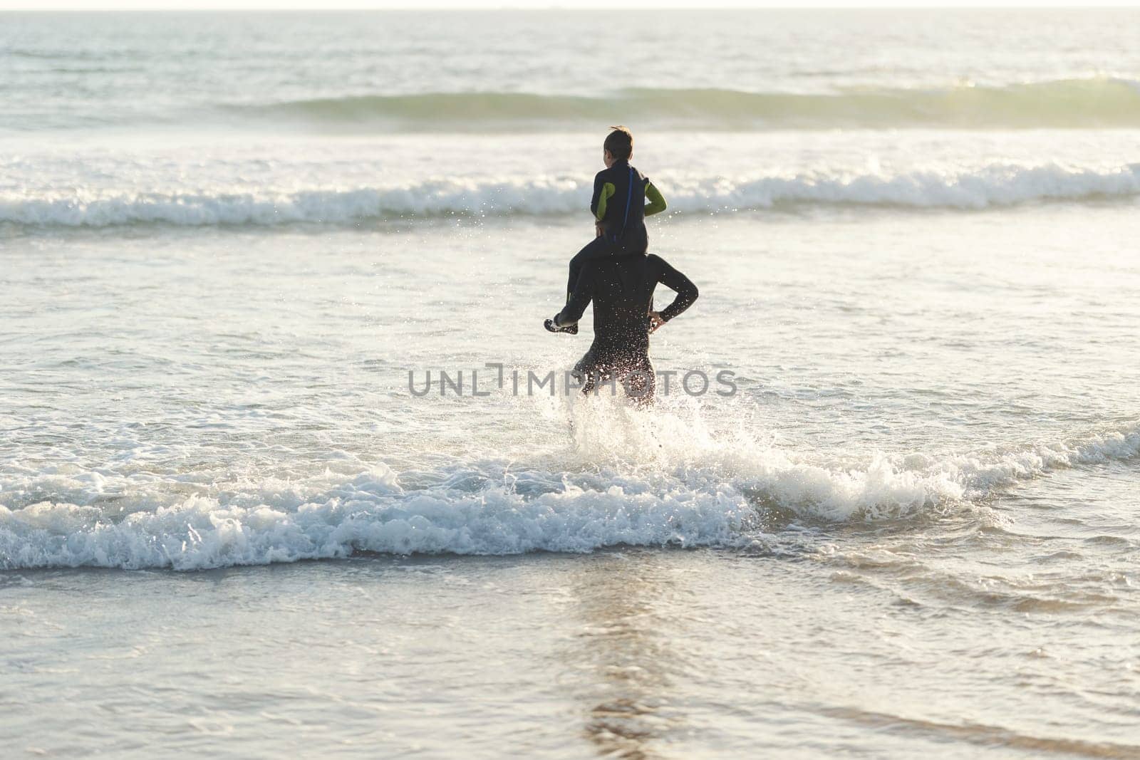 Little son on the shoulders of his father running to the sea at sunset lighting by Studia72