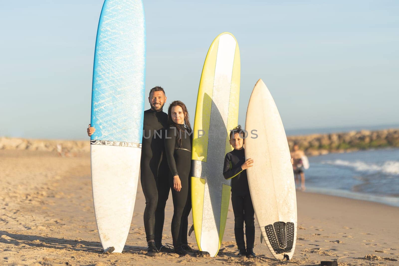 A family of surfers standing on the seashore in wetsuits by Studia72