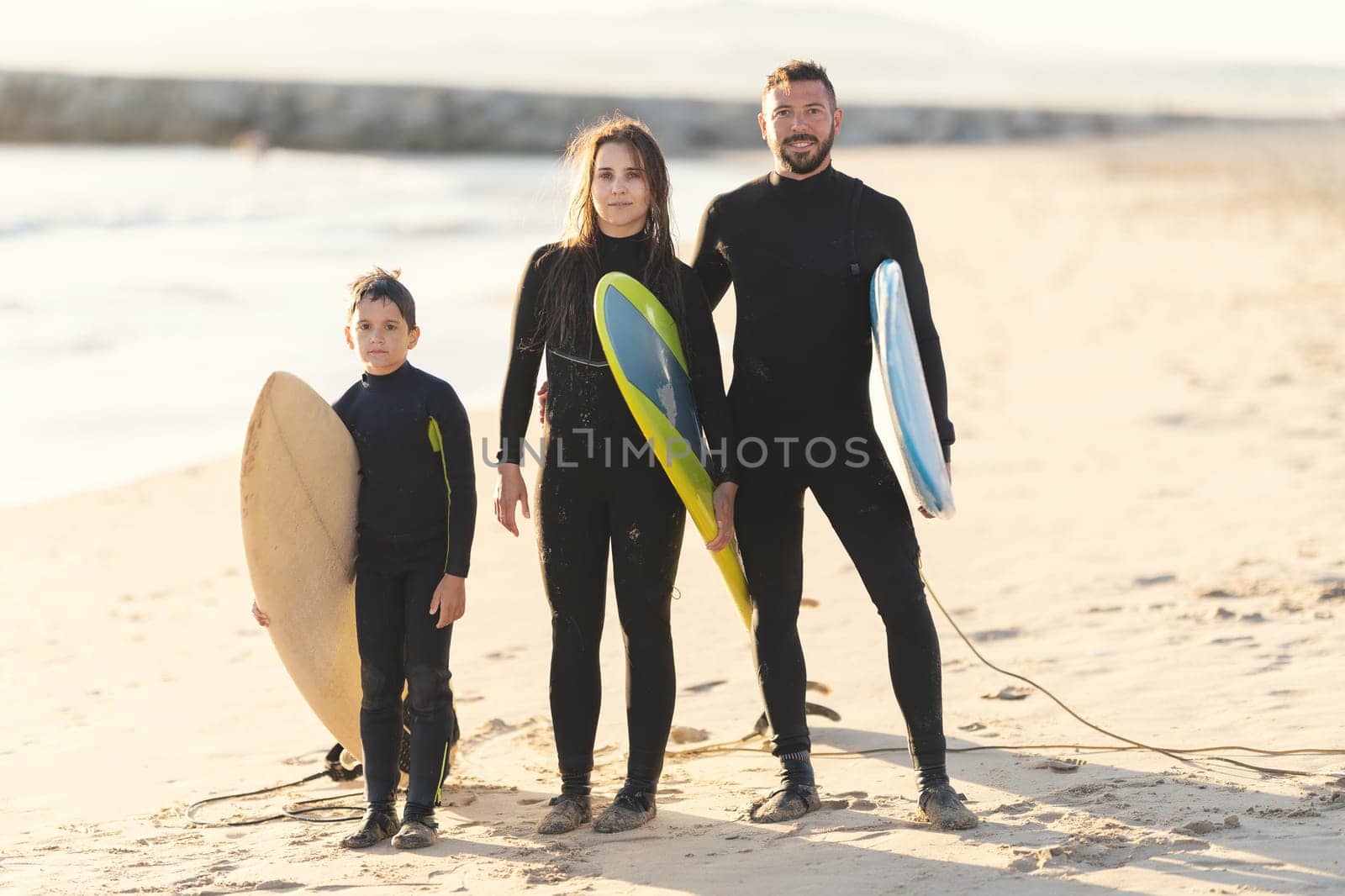 A family of surfers in wetsuits standing on the seashore holding surfboards. Mid shot