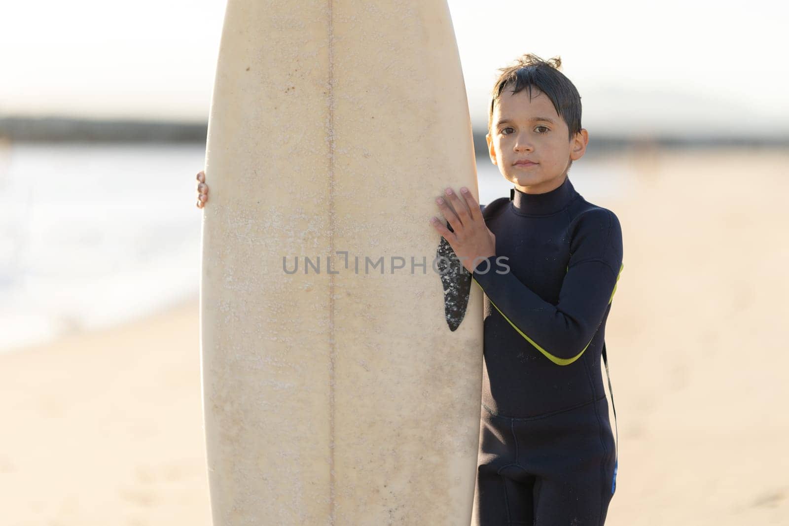 A little boy in a wetsuit holding a surfboard on the seashore. Mid shot
