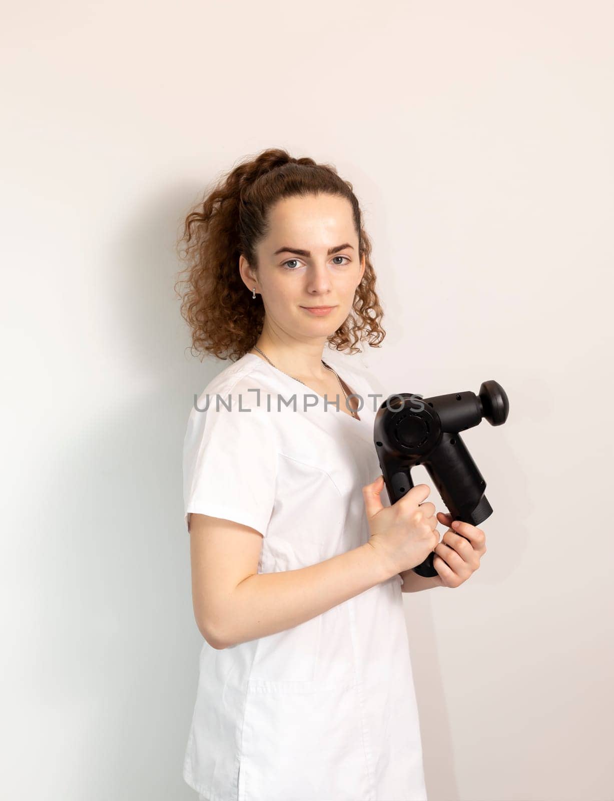 Portrait Caucasian Rehabilitation Specialist, Physician In White Medical Clothes Holds Massage Gun, Massager Product. Health Specialist, Rehabilitation. Vertical plane. High quality photo