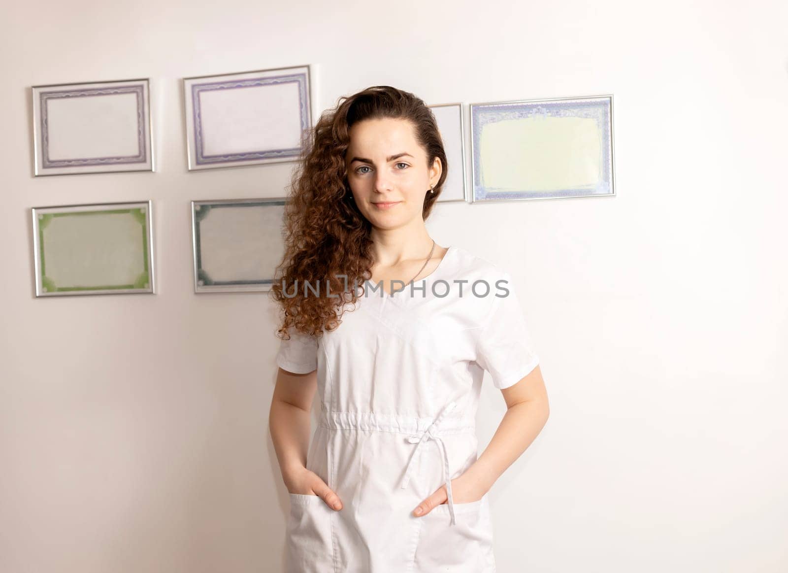 Portrait Smiling Young Female Rehabilitation Specialist, Physiotherapist In White Medical Clothes, Certificates In Frames On Wall On Background. Health Specialist, Rehabilitation. Horizontal plane.