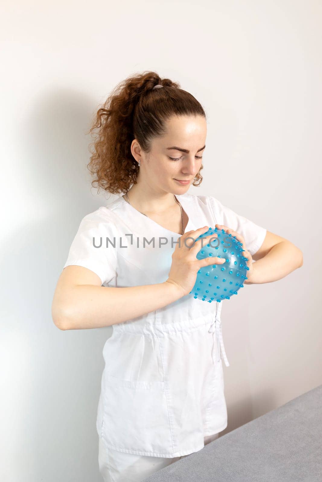 Caucasian Rehabilitation Specialist, Physiotherapist In White Medical Clothes Holds Massage Ball With Pimples In Therapeutic Cabinet. Health Specialist, Rehabilitation. Vertical. High quality photo