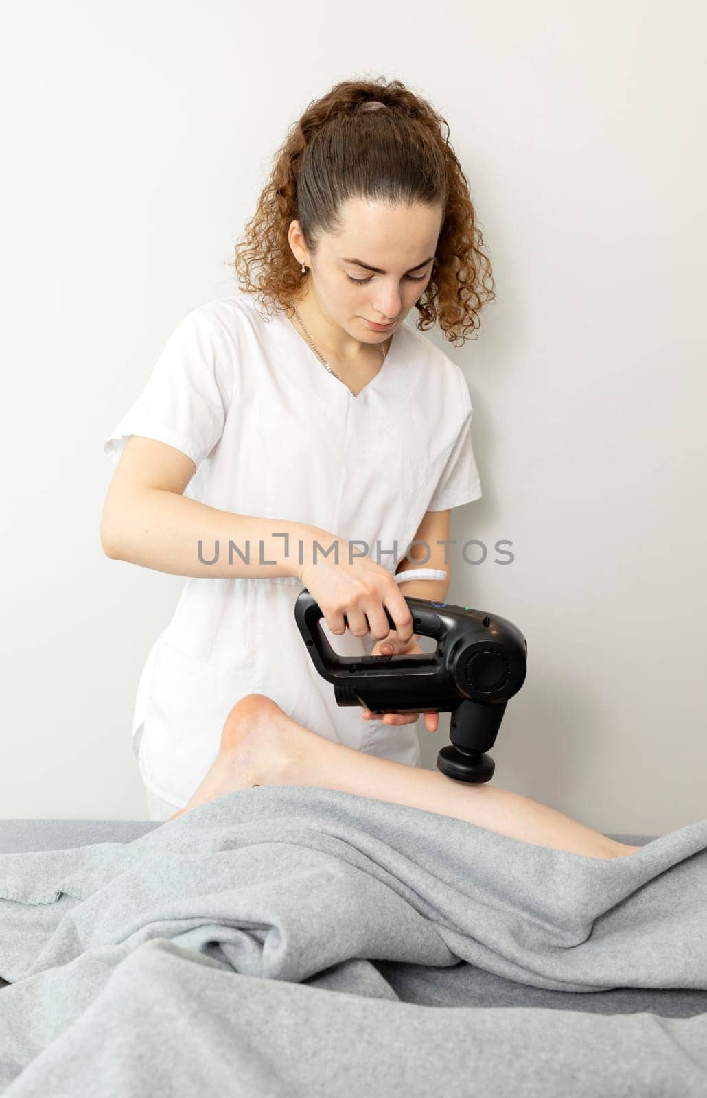 Pediatrician, Physical Therapist In White Medical Clothes Makes Leg Massage With Massager Product, Gun To Child. Health Specialist, Rehabilitation, Healthcare. Vertical plane. High quality photo
