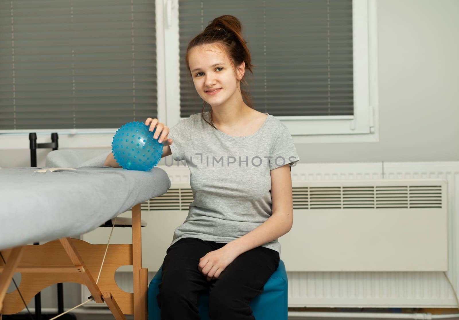 Portrait Of Cheerful Girl With Cerebral Palsy Sitting On Chair, Holding Blue Massage Ball With Pimples in Hospital. Rehabilitation. Balance And Posture Maintenance, Cure. Horizontal plane