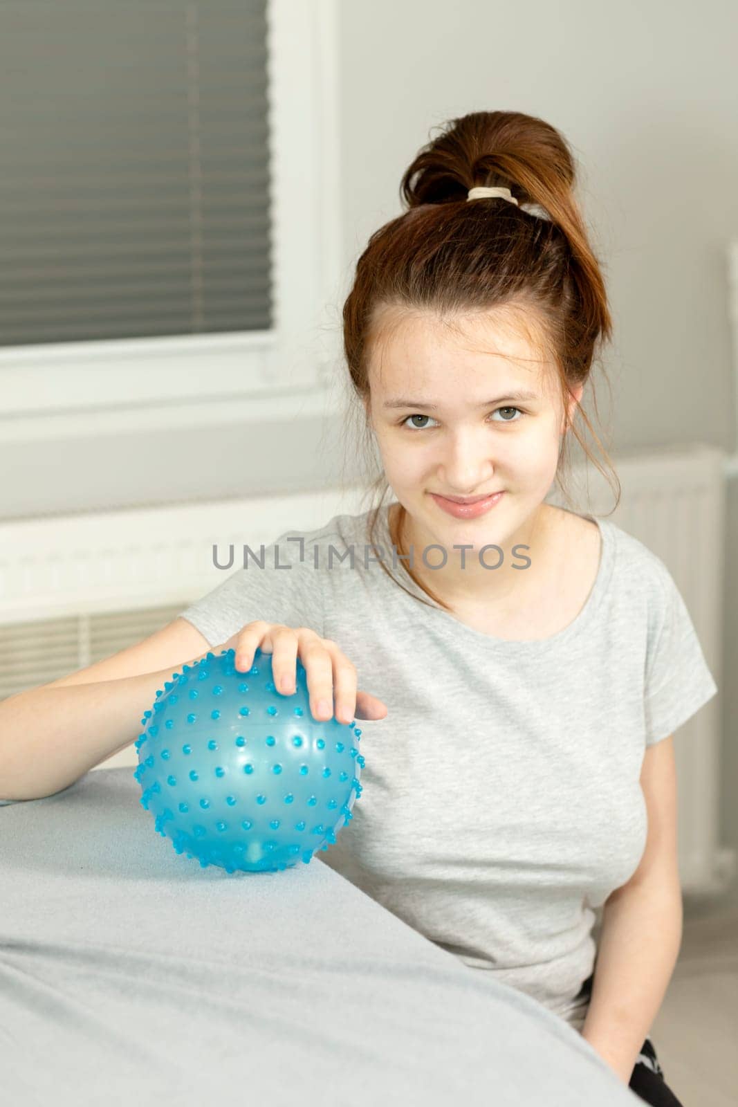 Portrait Of Cheerful Girl With Cerebral Palsy Sitting in Chair, Holding Blue Massage Ball with Pimples in Hospital. Rehabilitation. Balance And Posture Maintenance, Cure. Vertical plane.