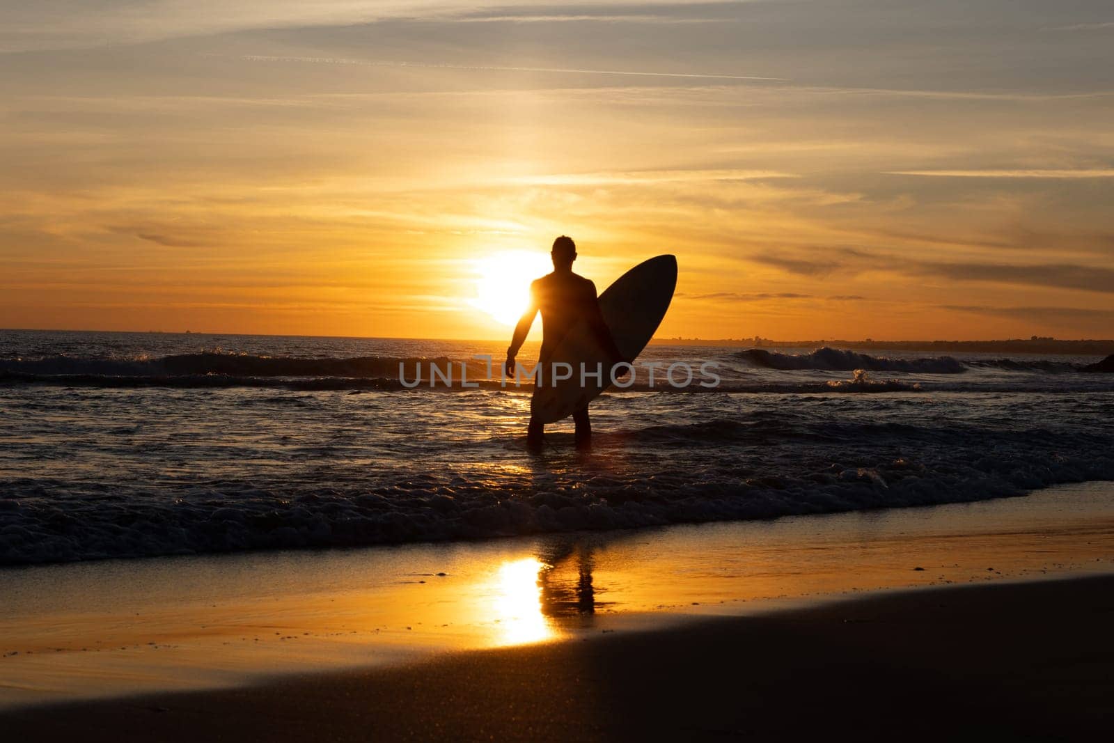 Silhouette of a man holding surfboard standing on the seashore at sunset. Mid shot