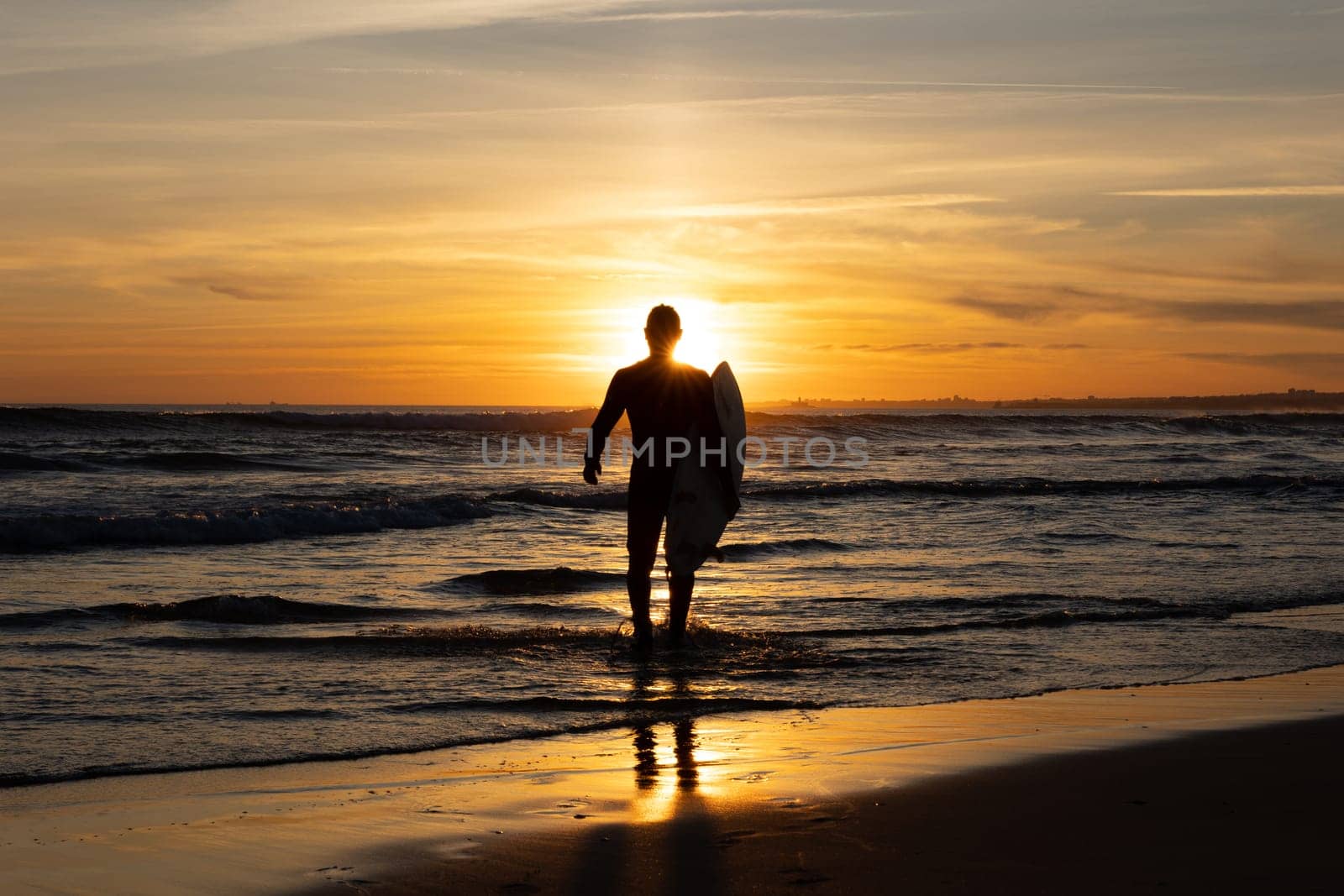 Dark silhouette of a man surfer walking out on the seashore holding a surfboard at bright sunset. Mid shot