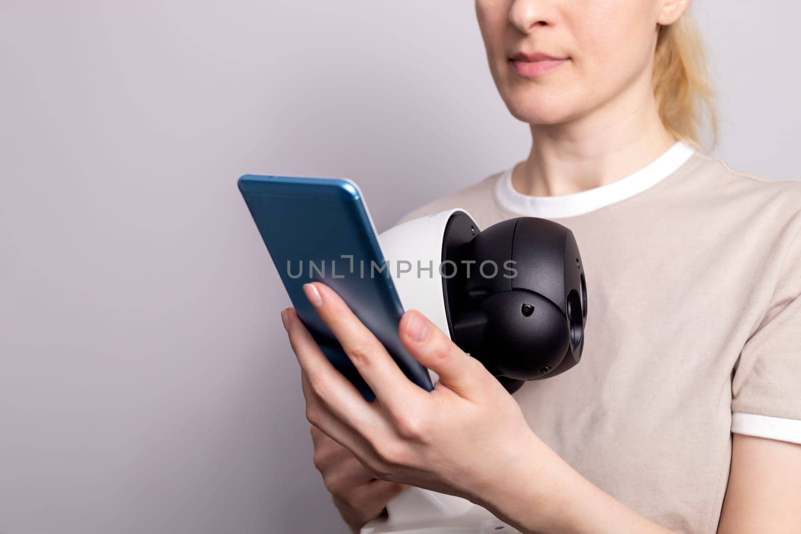 White Female Person Holds IP Wifi Outdoor Security Camera And Looks at Smartphone On White Grey Background. Smart Home, House Surveillance, Protection. Horizontal, closeup.