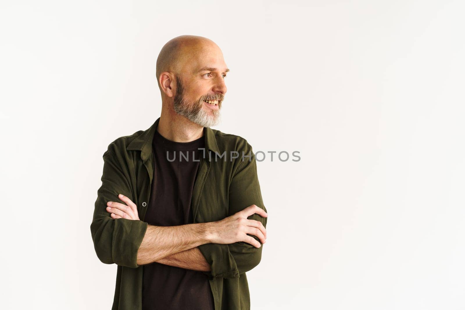 Senior man with warm smile, standing against white wall background, and crossing hands. Essence of happy old age concept, radiating positivity and joy. by LipikStockMedia