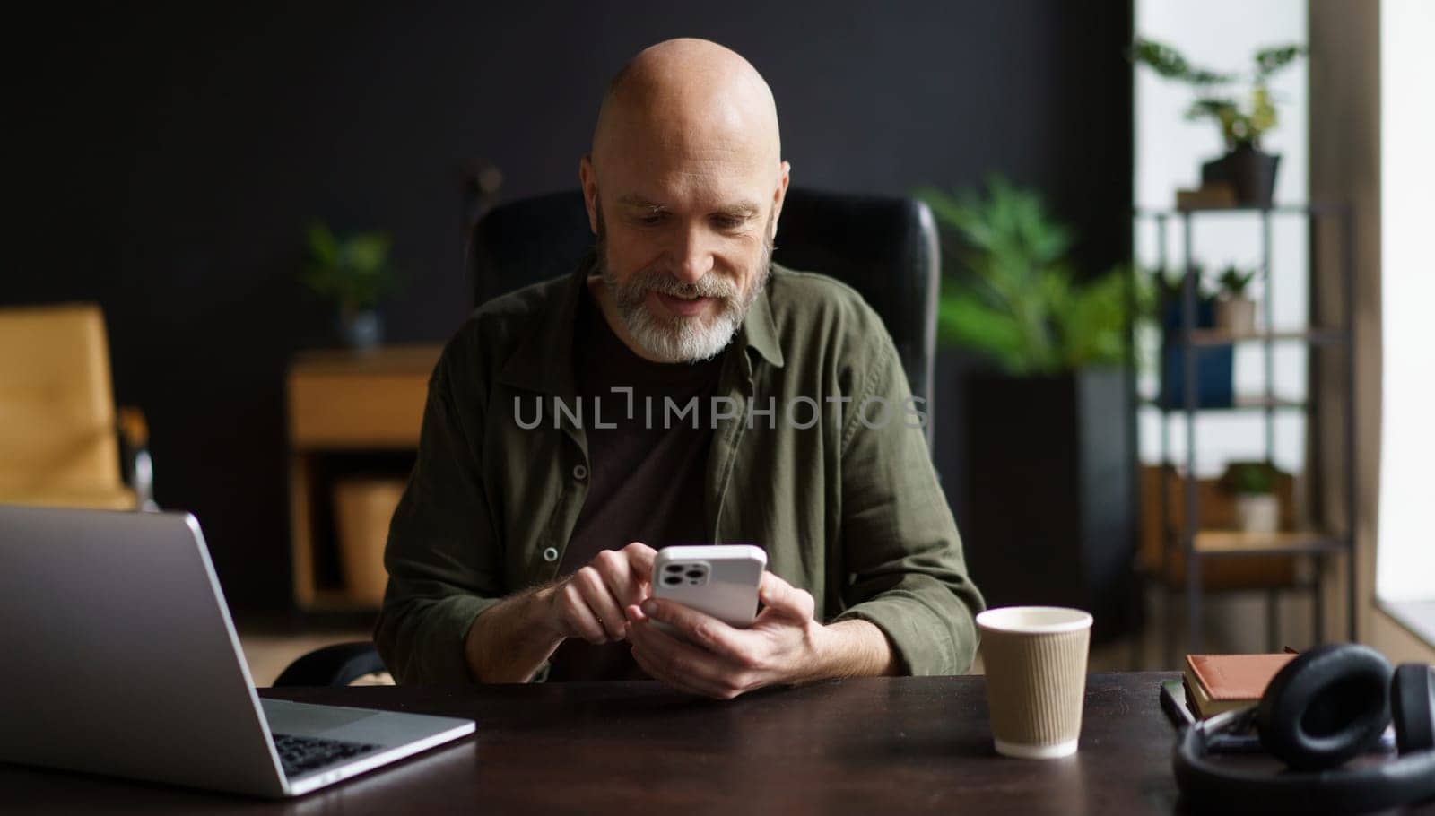Happy and charismatic senior man who engrossed in text message on phone while taking break from work on laptop. Senior man, full of vitality, seen enjoying comfort of own house. by LipikStockMedia