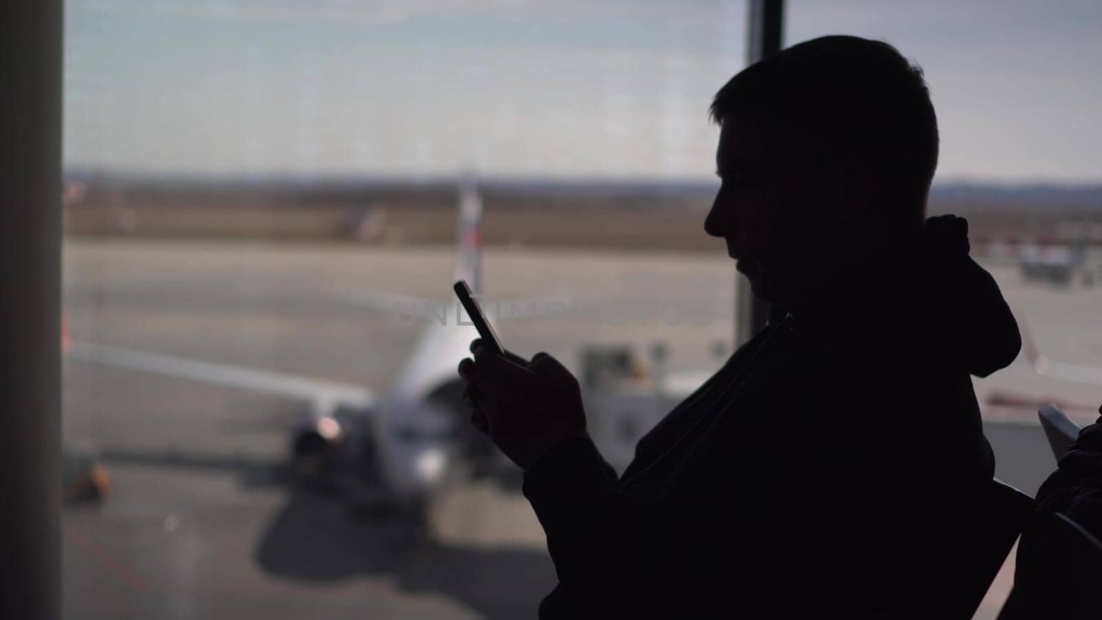 A young man in a sweatshirt is sitting at the airport with a phone in his hands. The man is using a smartphone. Silhouette of a man on the background of the planes outside the window. 4k