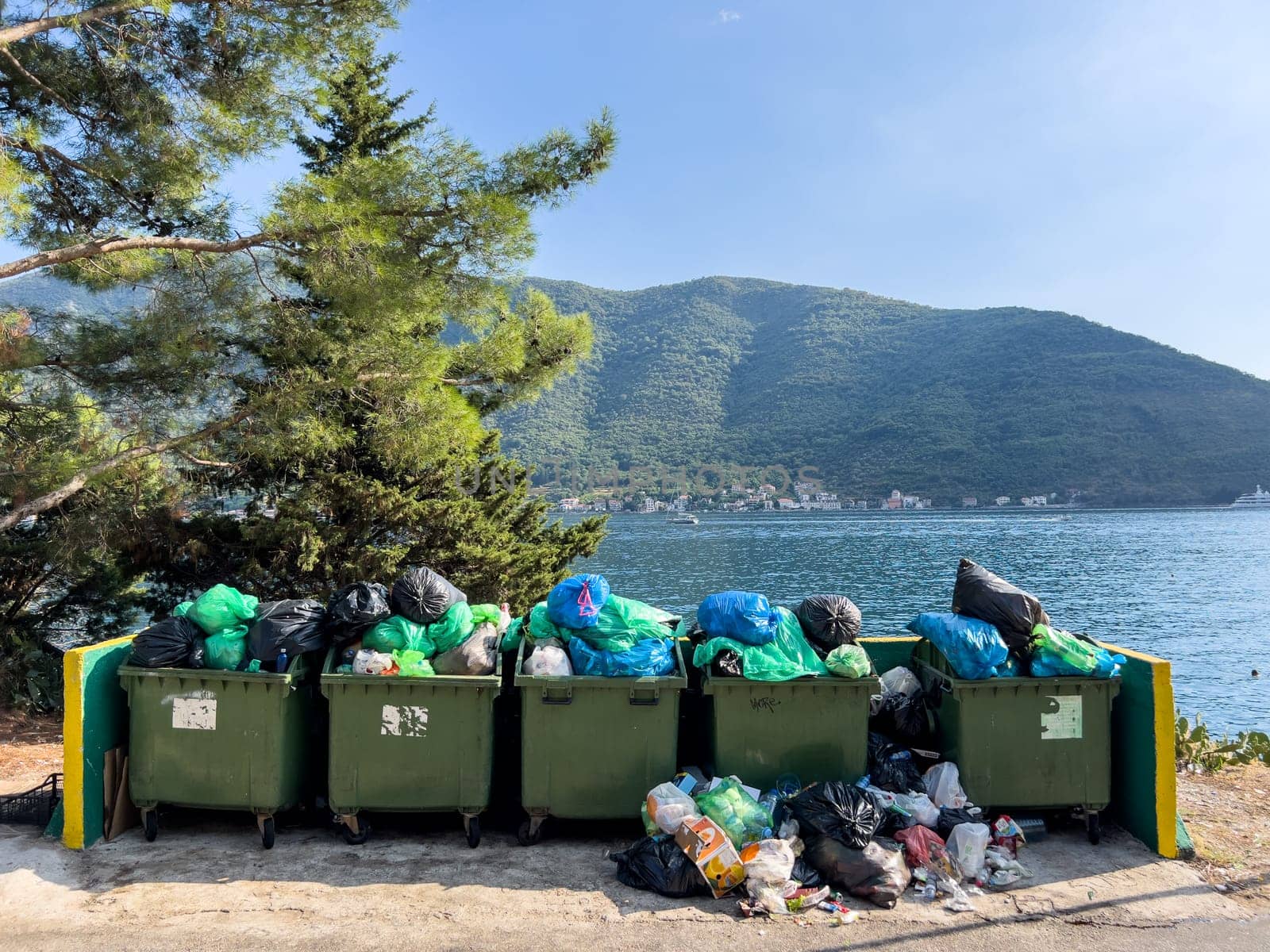 Overflowing garbage cans stand in a fence on the seashore near a tree. High quality photo