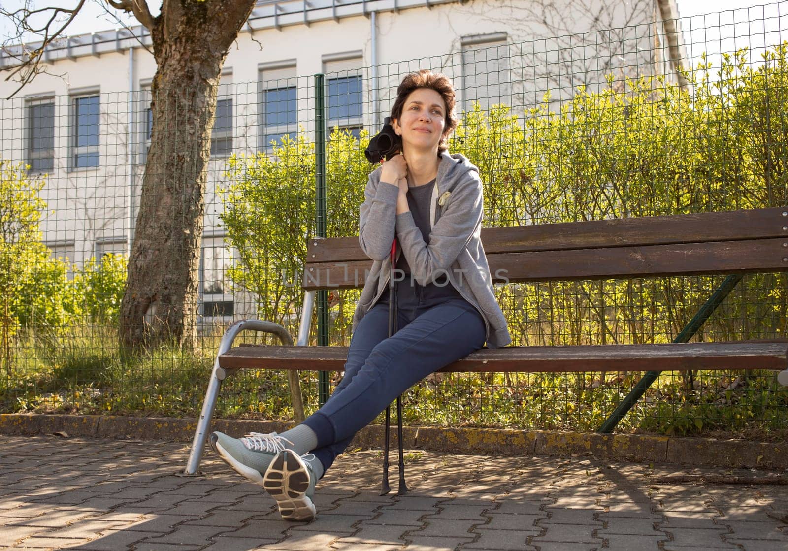 Pretty Woman Resting on Bench Holding Poles After Nordic Walking. Beautiful Happy Caucasian Woman Enjoys Sunny Day. Physical Activity With Walking Poles. Sports Tourism, Outdoor Recreation.Horizontal