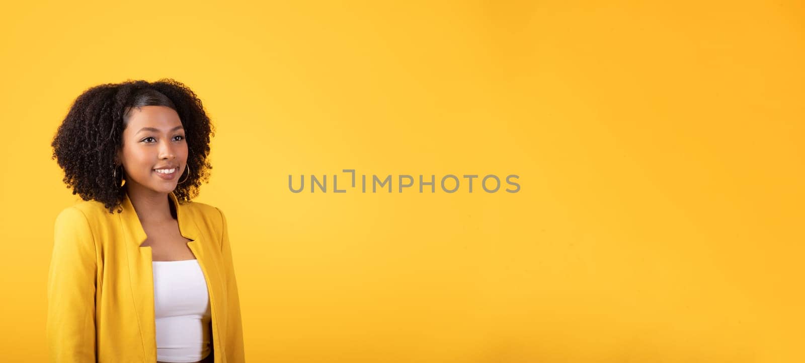 Pretty young african american curly woman looking at empty space and smiling, posing isolated on yellow studio background. Huge sale, good news, fashion lifestyle and beauty care