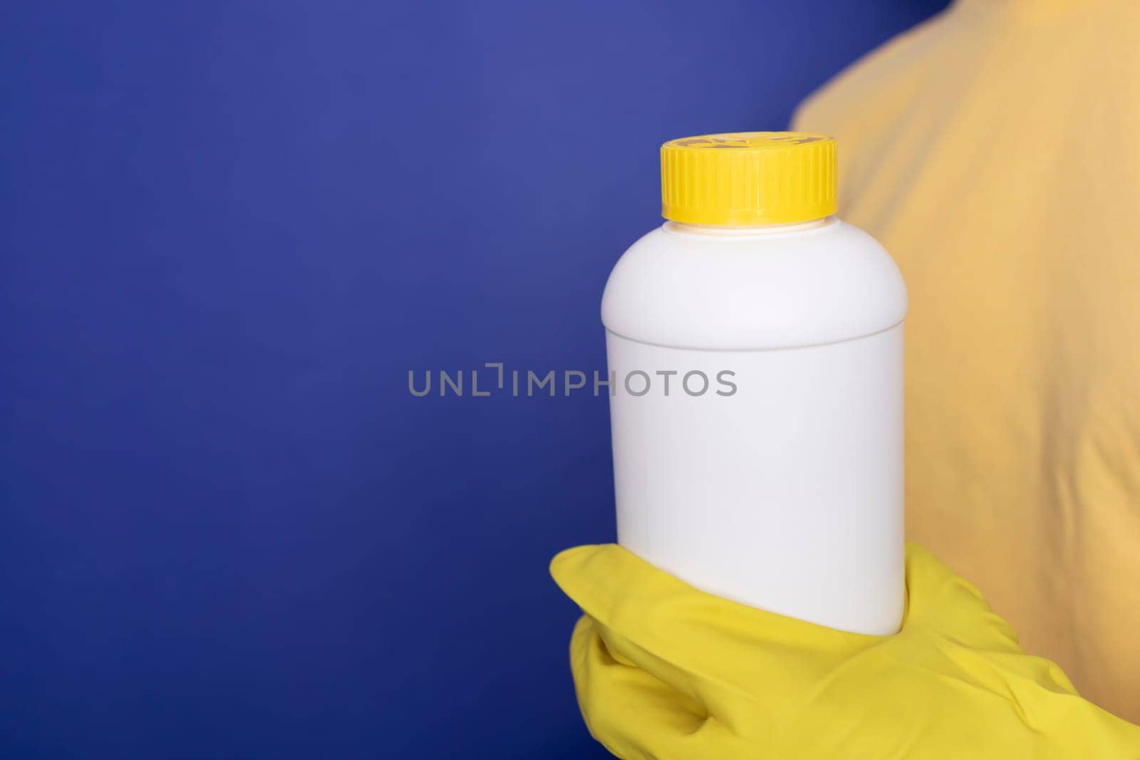 Woman Hand In Yellow Gloves Holds White Bottle With Pipe Cleaner Granules. Removal Of Blockage In Sink, Shower With Special Chemicals. Cleaning Clog In Kitchen, Bathroom. Blue On Background,Copyspace