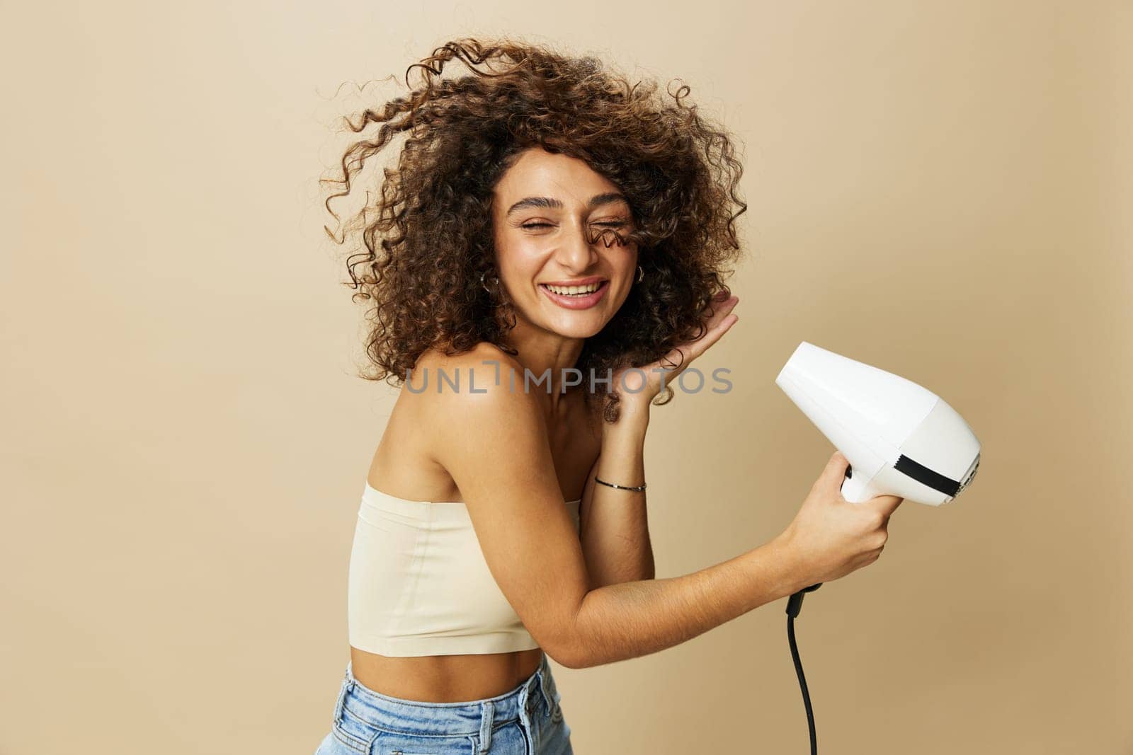 Woman dries curly afro hair with blow dryer, home beauty care styling products hair, smile on beige background by SHOTPRIME