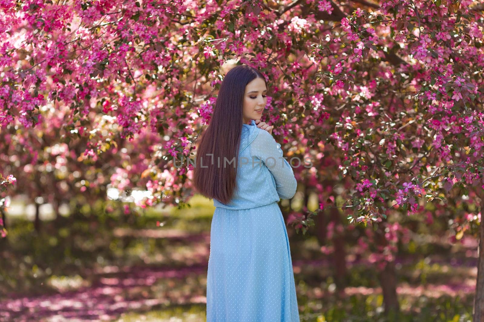 A pretty girl with long hair, in a light blue dress standing near pink blooming garden, in the garden. Rear view. Close up. Copy space