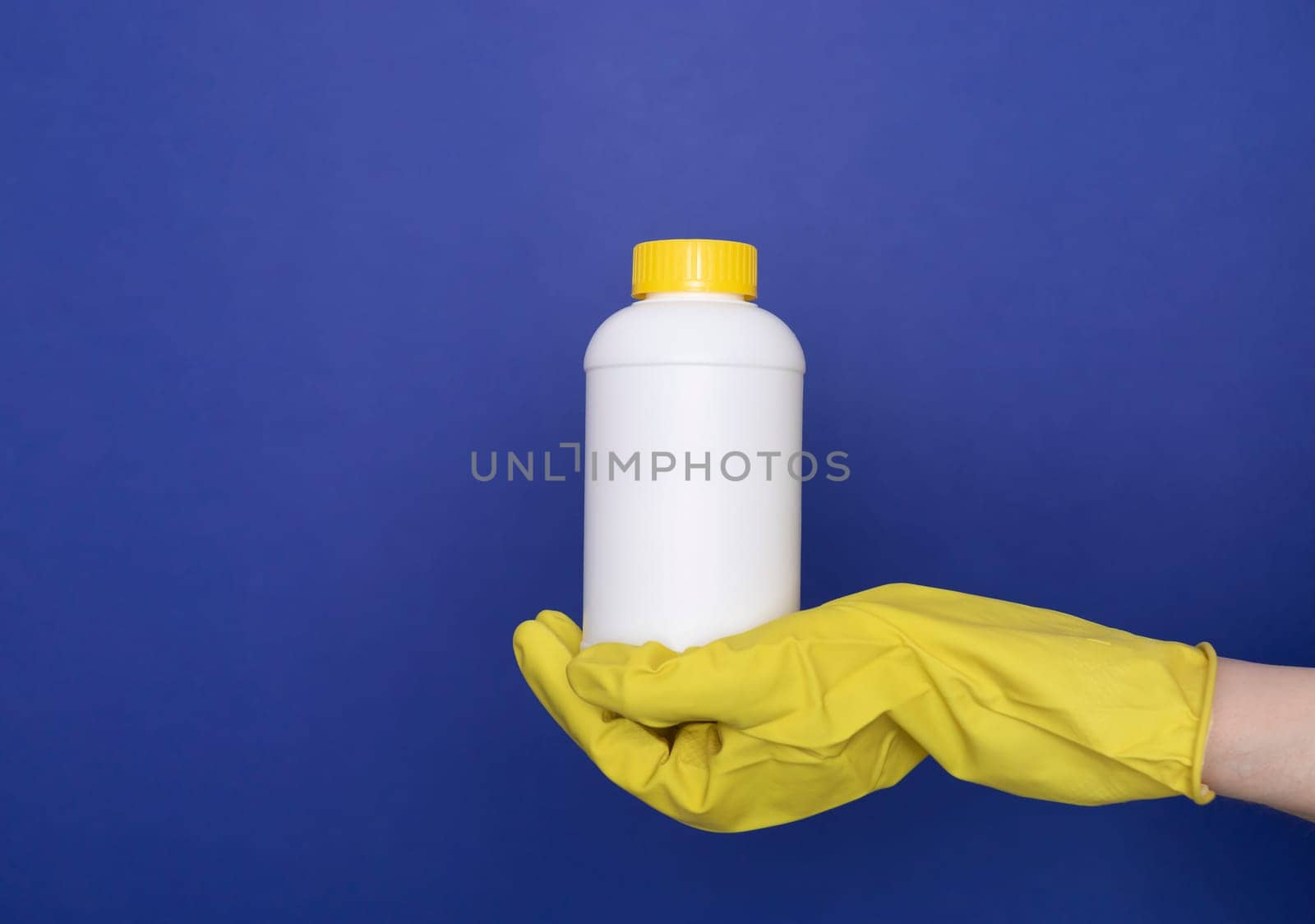 Human Hand In Yellow Gloves Holds White Bottle With Pipe Cleaner Granules. Removal Of Blockage In Sink, Shower With Special Chemicals. Cleaning Clog In Kitchen, Bathroom. Blue On Background, Copyspace