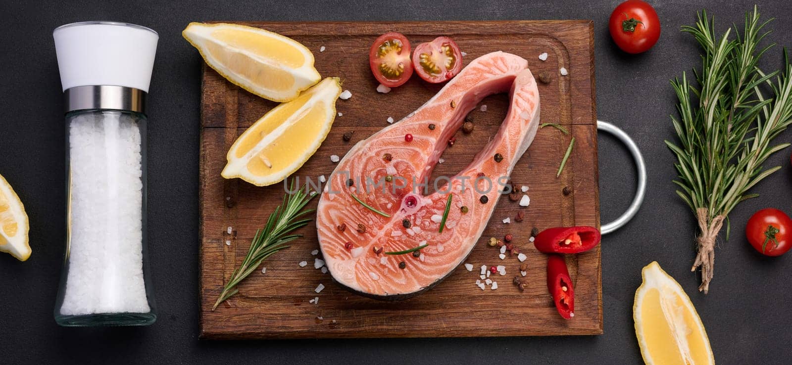 Raw salmon steak on a wooden cutting board, lemon slices, spices. Top view on black table