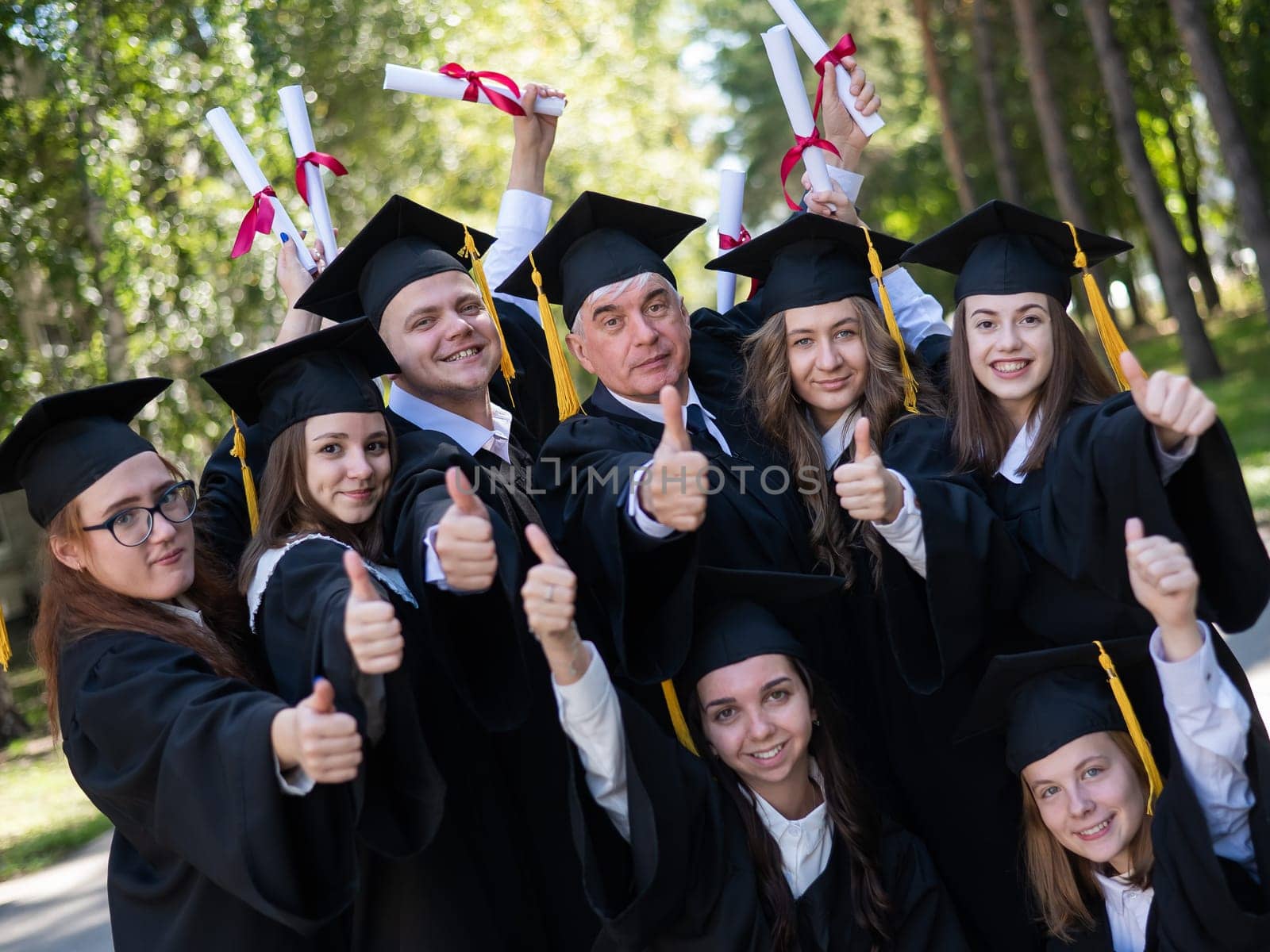 A group of graduates in robes give a thumbs up outdoors. Elderly student. by mrwed54