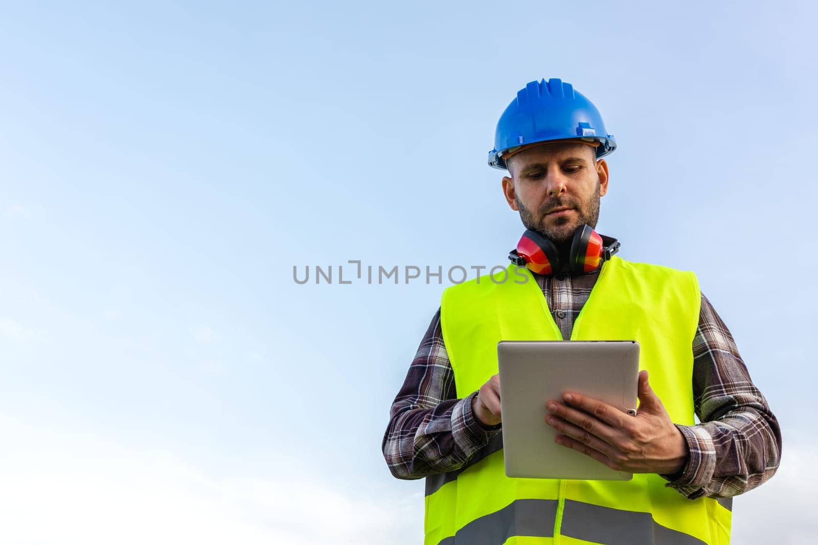 Man wearing helmet and vest checking information on digital tablet outdoors. Blue sky, copy space. by Hoverstock