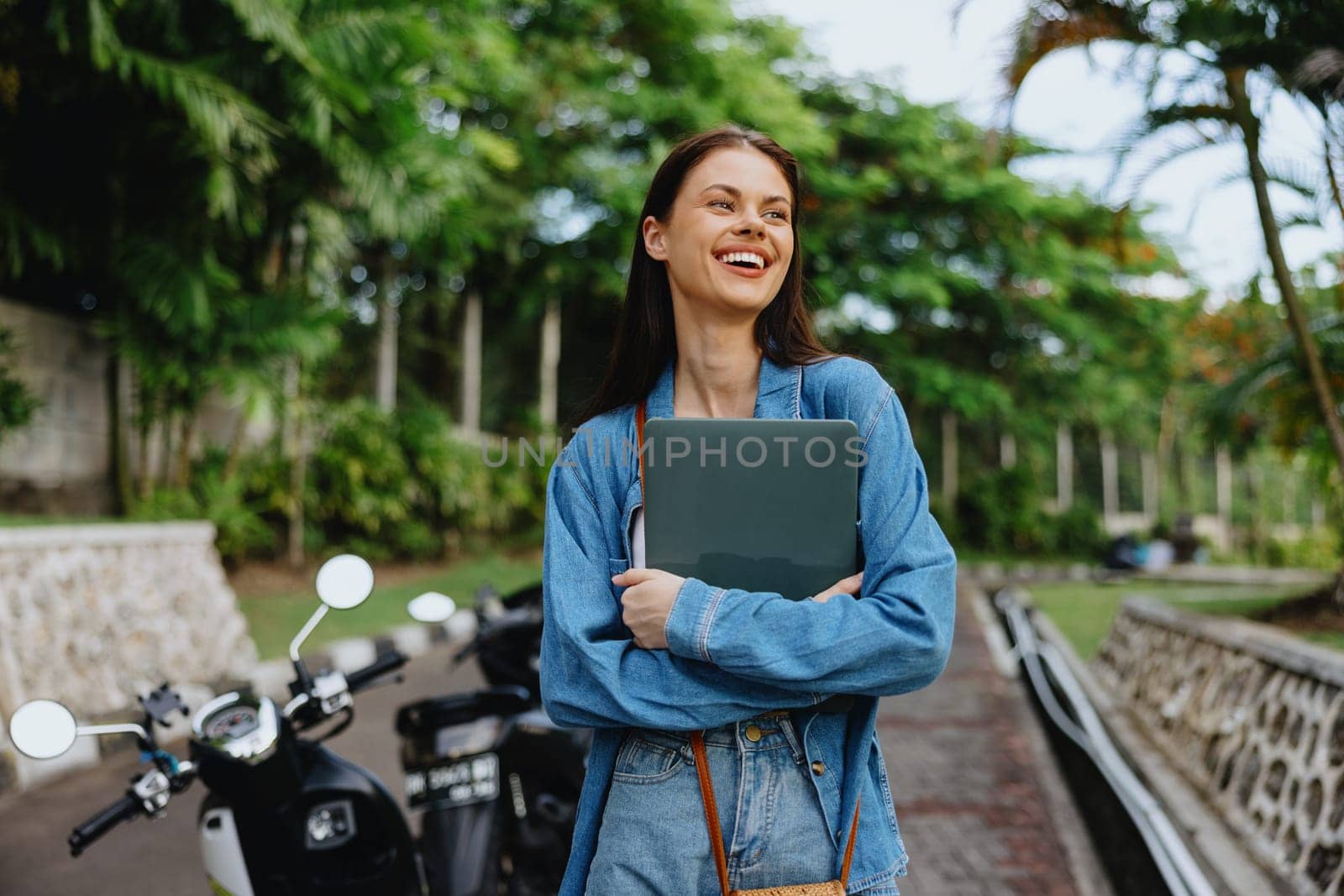 Woman smiling walking in the park outside with laptop freelancer against a backdrop of green palm trees in summer, tropical backdrop, blogger on a trip, work online. High quality photo
