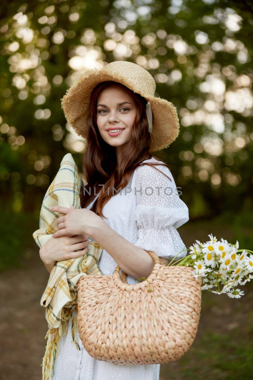 a woman with a wicker hat, a bag and a plaid smiles while standing in nature in the park by Vichizh