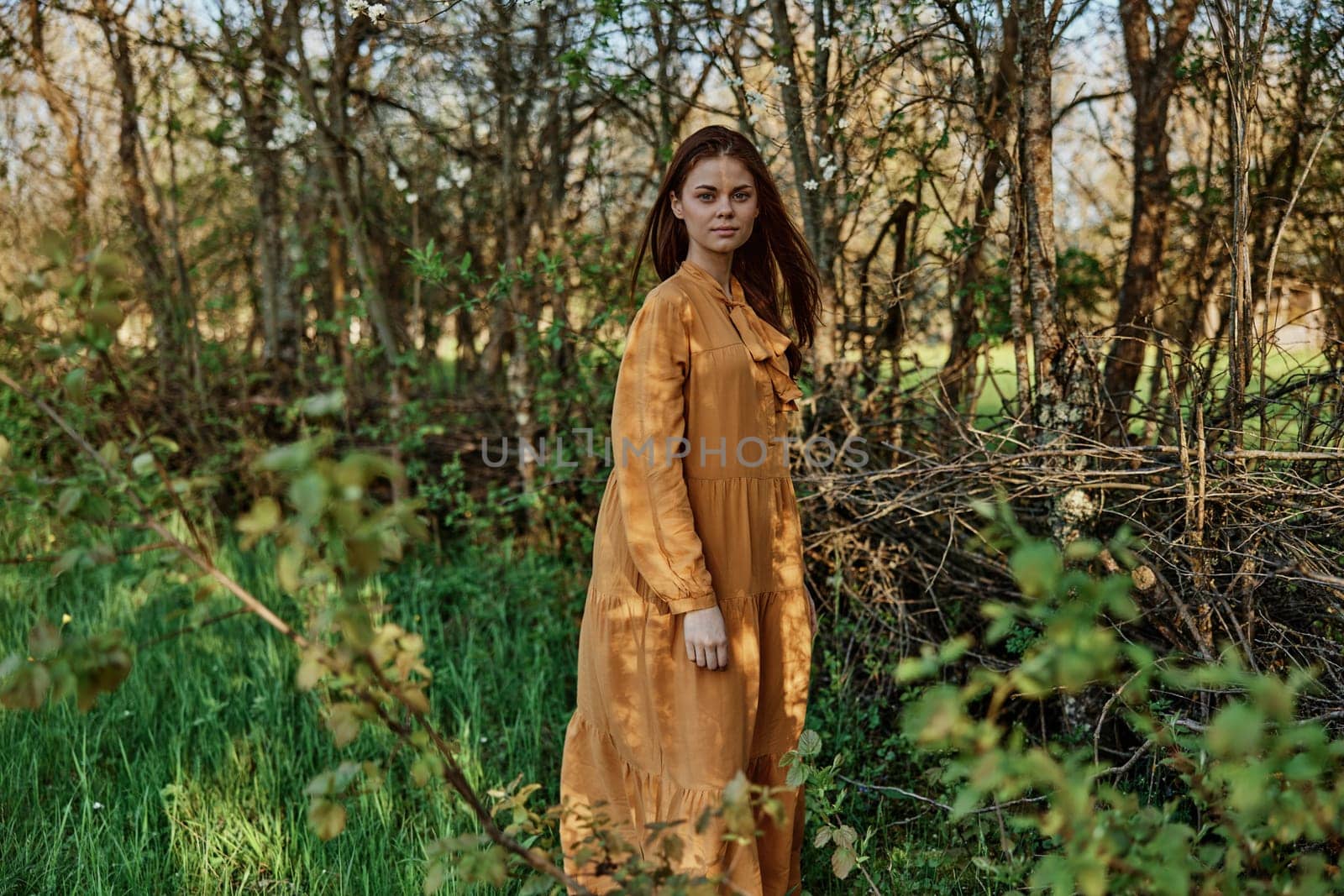 a woman with long hair walks in the shade near the trees, dressed in a long orange dress, enjoying the weather and the weekend, while looking at the camera. The theme of privacy with nature by Vichizh