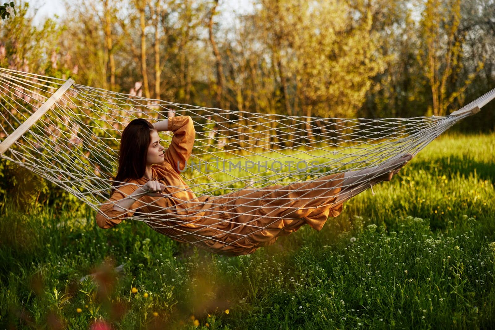 an elegant, slender woman is resting in nature lying in a mesh hammock in a long orange dress enjoying the rays of the setting sun on a warm summer day looking away by Vichizh