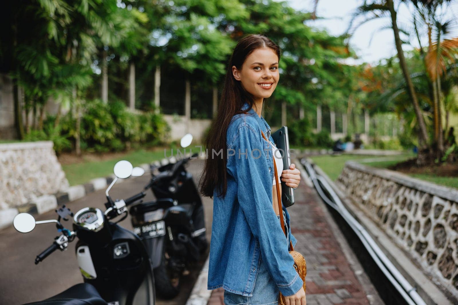 Woman smiling walking in the park outside with laptop freelancer against a backdrop of green palm trees in summer, tropical backdrop, blogger on a trip, work online by SHOTPRIME