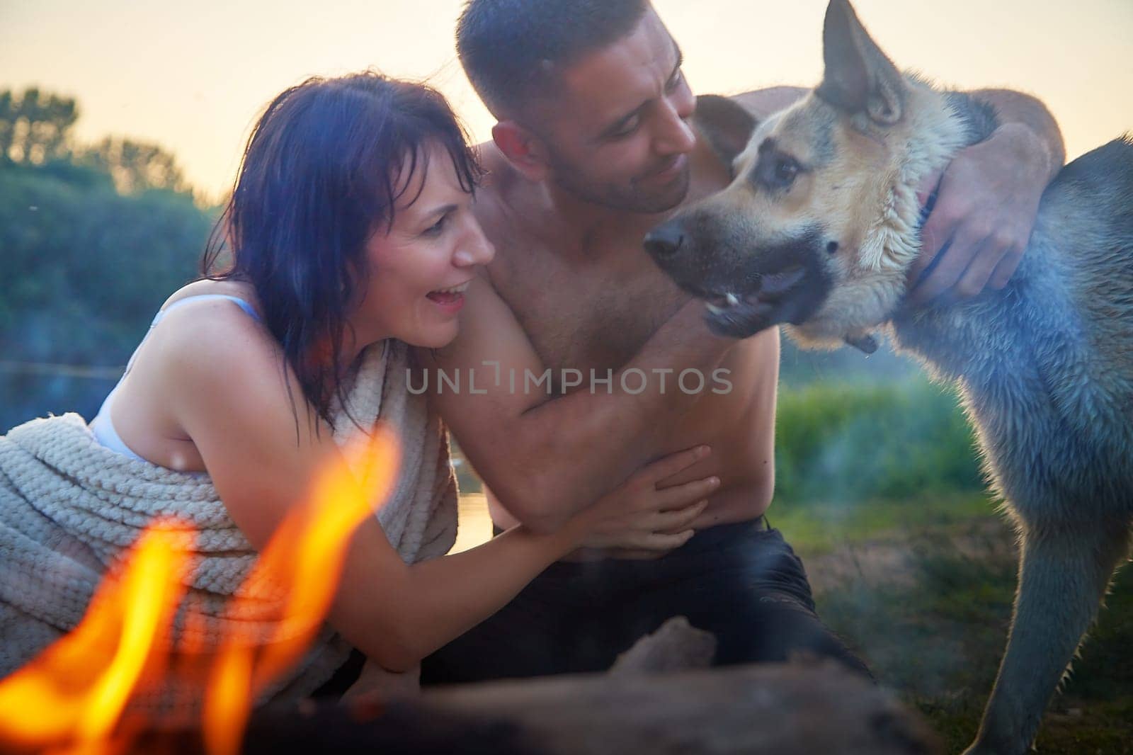 Happy wet couple relaxin, having fun and hugs with big dog near fire in camping on nature at summer sunny evening in sunset. Family or lovers have date and rest outdoor. Concept of love by keleny
