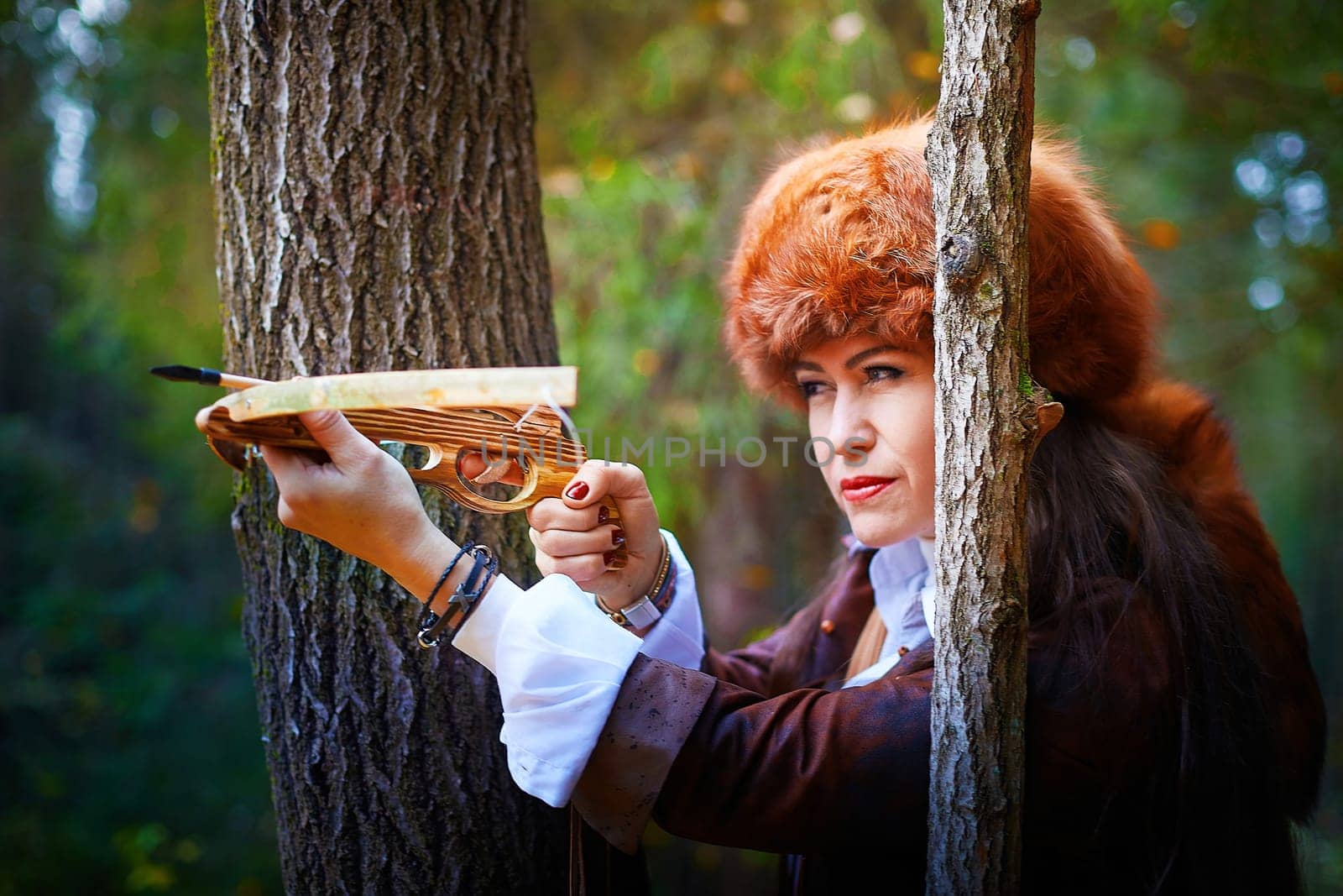 Girl in a leather jacket, a big red fox fur hat and with a crossbow in the forest in autumn. A female model poses as a fabulous royal huntress on nature hunt at a photo shoot